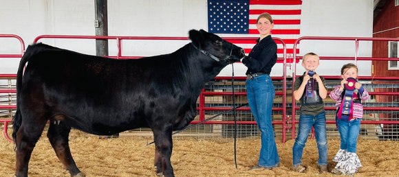 A show cow and owner posing next to two children holding up the award ribbons.